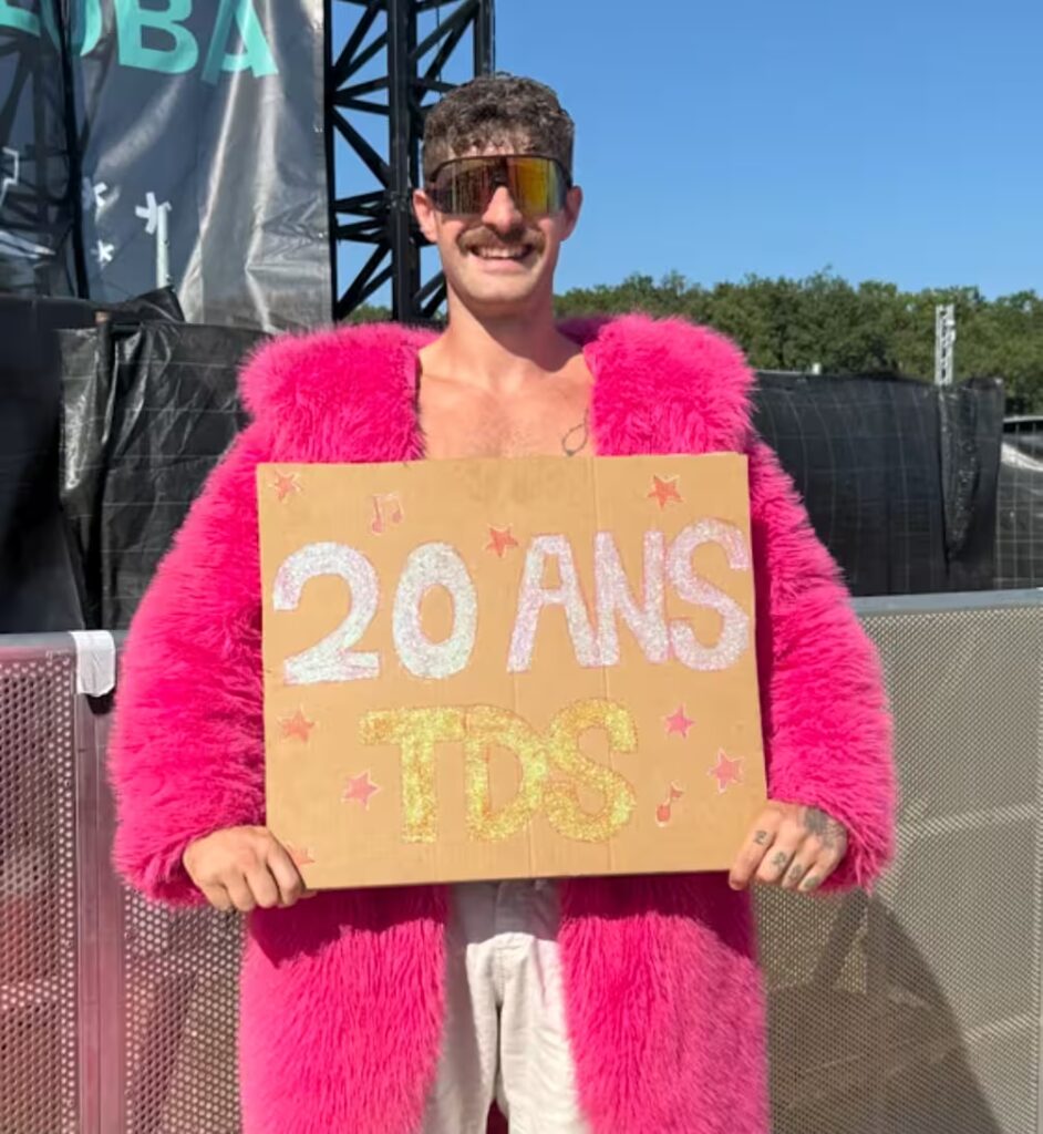 Un festivalier tout de rose vêtu tient un panneau 20 ans en acrton